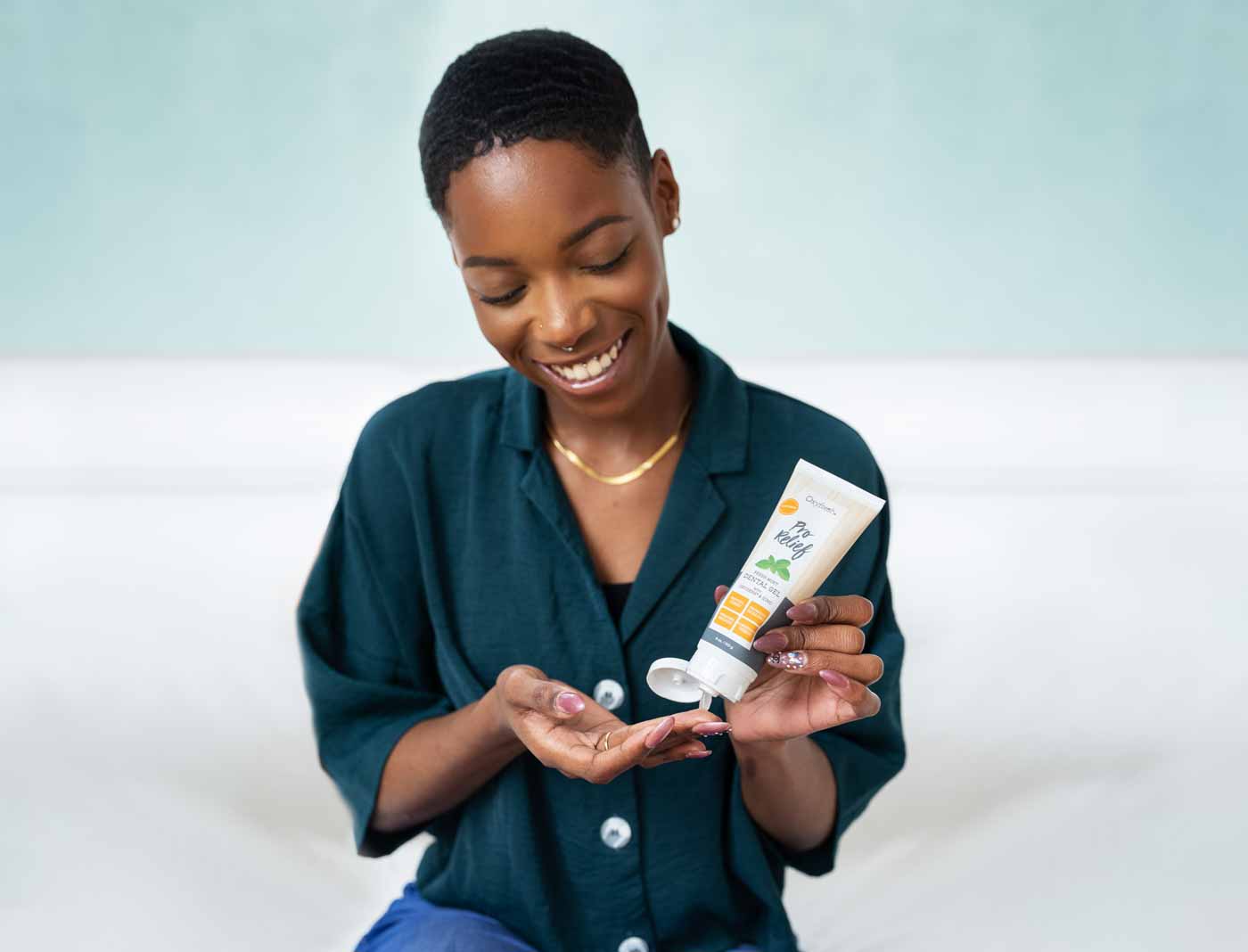 lady-with-short-cropped-hair-wearing-an-emerald-button-down-shirt-and-a-gold-chain-smiling-looking-down-at-the-oxyfresh-pro-relief-gel-that-she-is-placing-on-her-finger-for-application